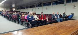 #DescriçãodaImagem a foto foi tirada no auditório onde aparecem varias pessoas sentadas, sendo uma delas cadeira e uma pessoa  segura uma bengala. 
